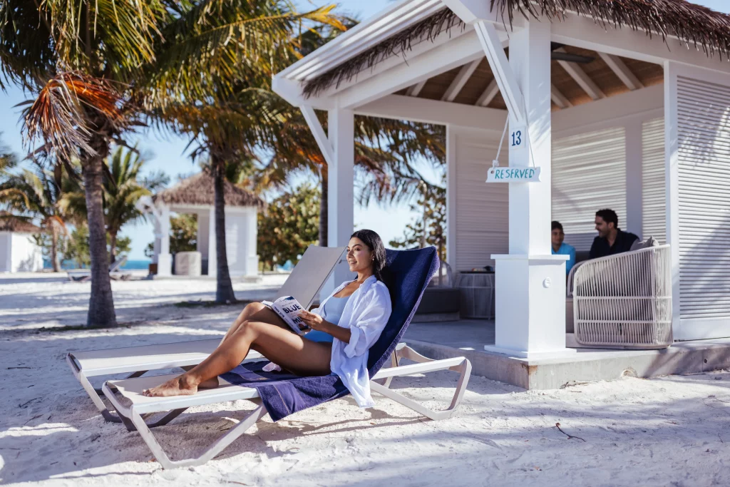 Eine Frau, die sich auf einem Liegestuhl entspannt und an einem Sandstrand in der Nähe einer weißen Cabana ein Buch liest, im Hintergrund Palmen und zwei Personen – eine idyllische Szene der Muße, die die künstlerische Qualität professioneller Bildproduktion unterstreicht.