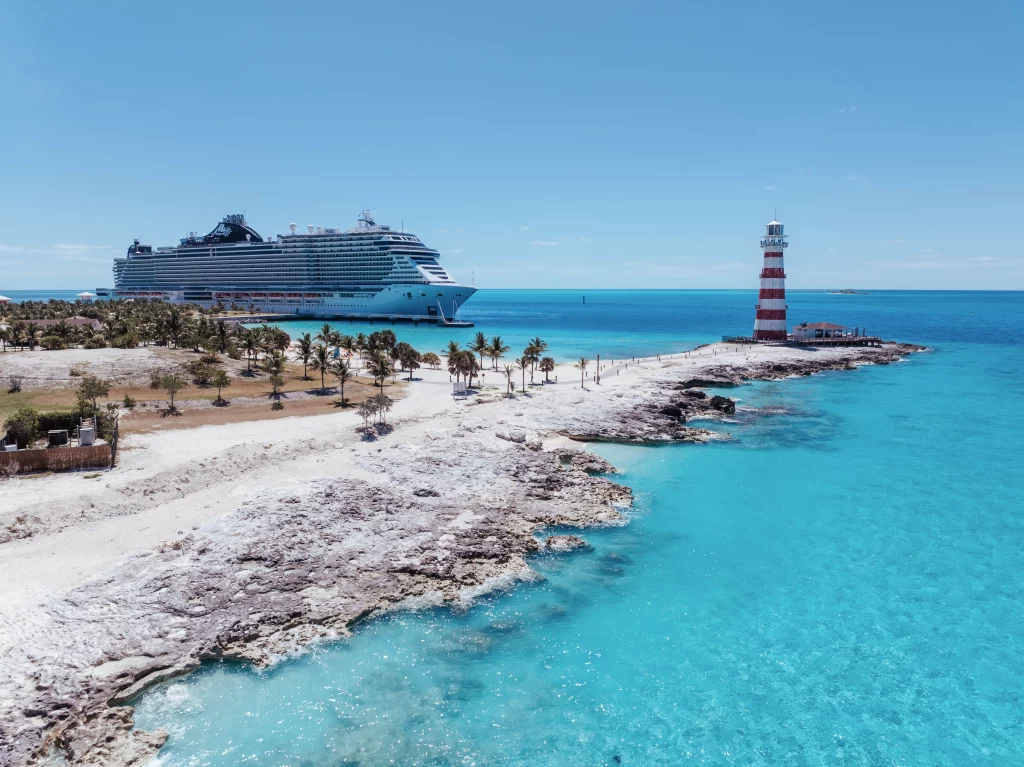 Ein großes Kreuzfahrtschiff liegt vor Anker in der Nähe einer tropischen Insel mit klarem, blauem Wasser, einer felsigen Küste, Palmen und einem rot-weiß gestreiften Leuchtturm – eine idyllische Kulisse, die sich perfekt für jede Bildproduktion eignet.