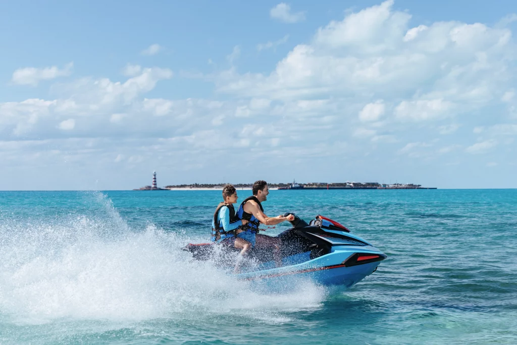 Zwei Personen in Schwimmwesten fahren auf einem blauen Jetski über türkisfarbenes Wasser – eingefangen im lebendigen Stil von Imageproduktion, im Hintergrund sind unter einem teilweise bewölkten Himmel eine Insel und ein Leuchtturm zu sehen.