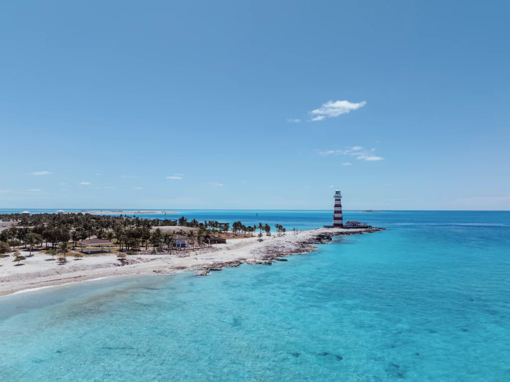Luftaufnahme einer Küstenlandschaft mit klarem, türkisfarbenem Wasser, einem Sandstrand, Palmen und einem rot-weiß gestreiften Leuchtturm unter strahlend blauem Himmel – ein Beispiel für herausragende Bildproduktion.