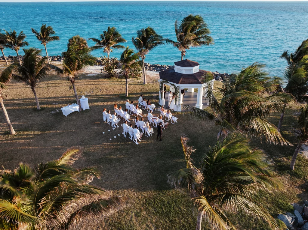 Eine kleine Hochzeitszeremonie im Freien findet am Meer unter einem Pavillon statt, umgeben von Palmen und Gästen, die auf weißen Stühlen sitzen – wunderschön festgehalten von der professionellen Bildproduktion.