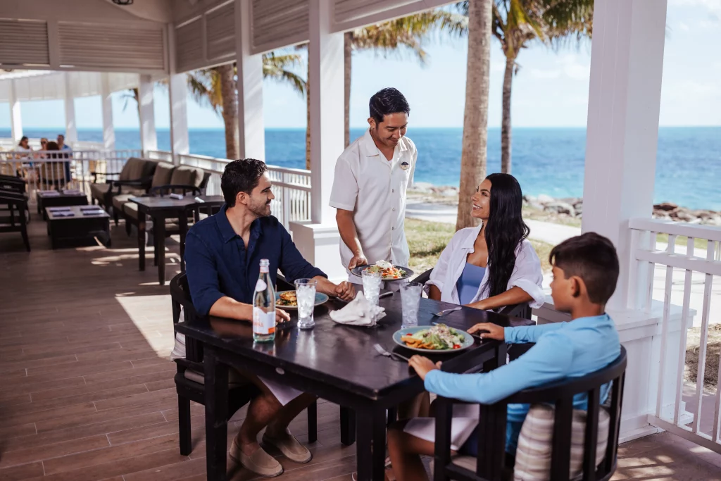 Ein Kellner bringt einer vierköpfigen Familie, die in einem Restaurant im Freien am Meer speist, das Essen – im Hintergrund sind Palmen und Terrassenmöbel zu sehen – eine perfekte Kulisse für eine Bildproduktion, die das entspannte Essen an der Küste einfängt.