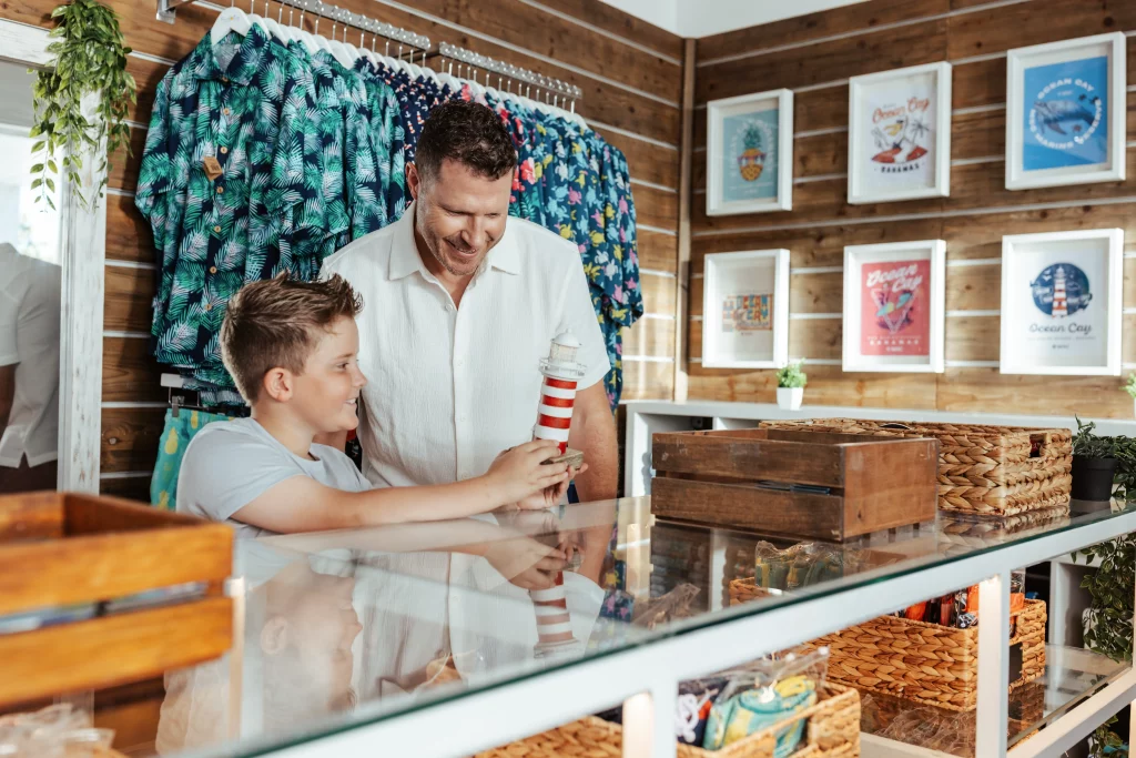 Ein Mann und ein Junge stehen an einer Ladentheke, lächeln und halten ein gestreiftes Leuchtturm-Modell in den Händen – ein Bild, das diesen fröhlichen Moment einfängt. An den Holzwänden hinter ihnen sind bunte Hemden und gerahmte Drucke ausgestellt.