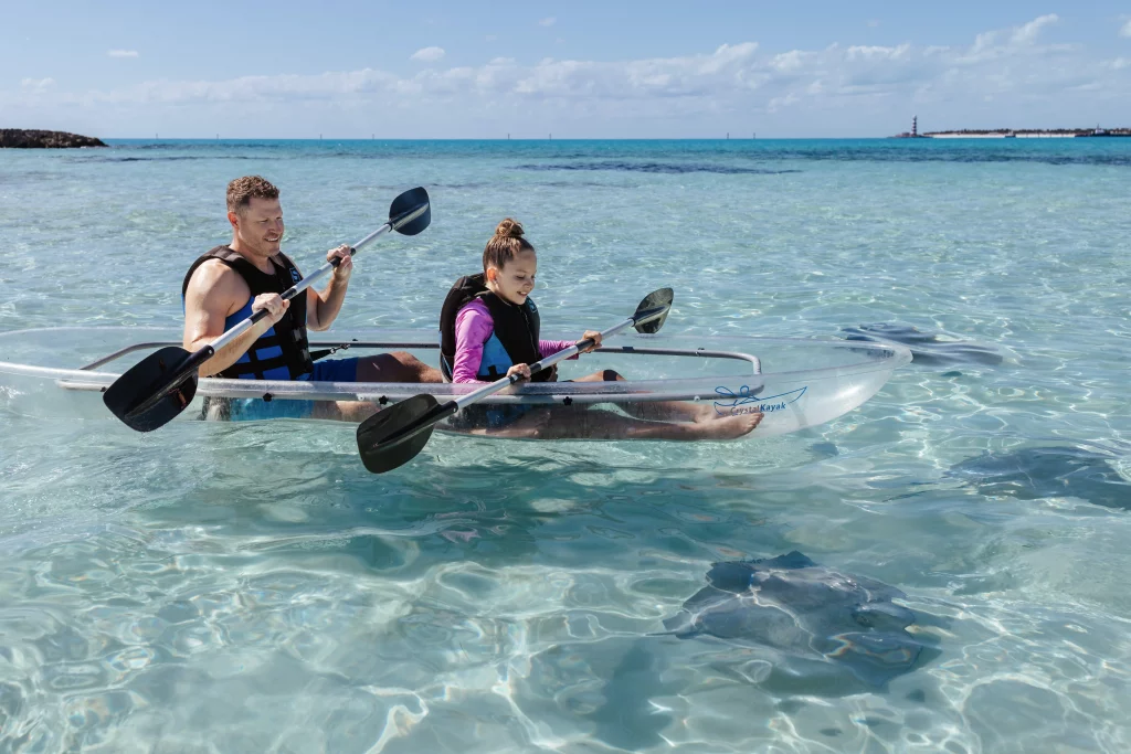 An einem sonnigen Tag paddeln zwei Personen in Schwimmwesten in einem durchsichtigen Kajak durch klares, flaches Wasser, unter dessen Oberfläche Stachelrochen zu sehen sind – eine atemberaubende Szene, die sich perfekt für die Bildproduktion eignet.