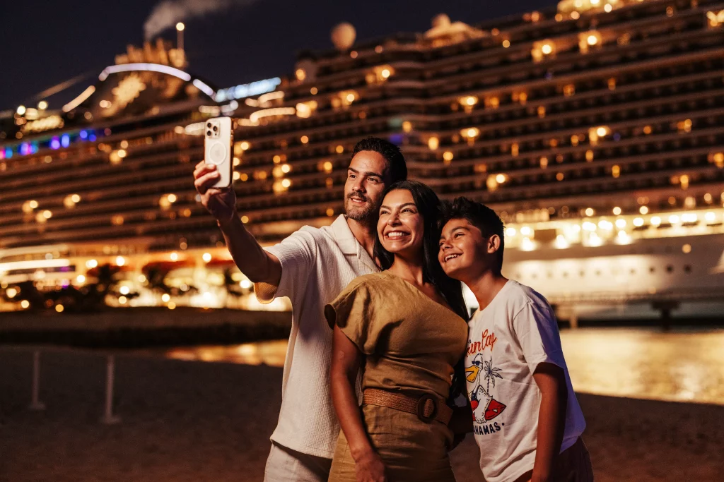Eine dreiköpfige Familie macht nachts am Strand ein Selfie mit einem hell erleuchteten Kreuzfahrtschiff im Hintergrund – ein Moment, der durch professionelle Bildproduktion perfekt eingefangen wurde.