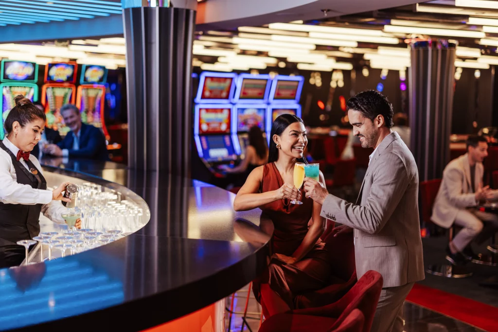 Eine Frau sitzt an der Bar eines Casinos an Bord der MSC World America und lächelt einen Mann im Anzug an, der Getränke in der Hand hält; ein Barkeeper mixt Cocktails, während Spielautomaten den Hintergrund erhellen – ein einladender Moment der Bildproduktion nach zwei Wochen auf See.