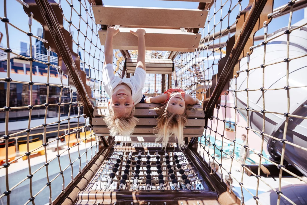 Zwei Kinder hängen kopfüber an einer Holzbrücke auf einem Spielplatz, lächeln in die Kamera, umgeben von Netzklettern und Spielgeräten an einem sonnigen Tag – eine fröhliche Szene, die an ein Abenteuer mit der MSC World America oder an Momente erinnert, die während einer Imageproduktion für „Zwei Wochen auf See“ festgehalten wurden.