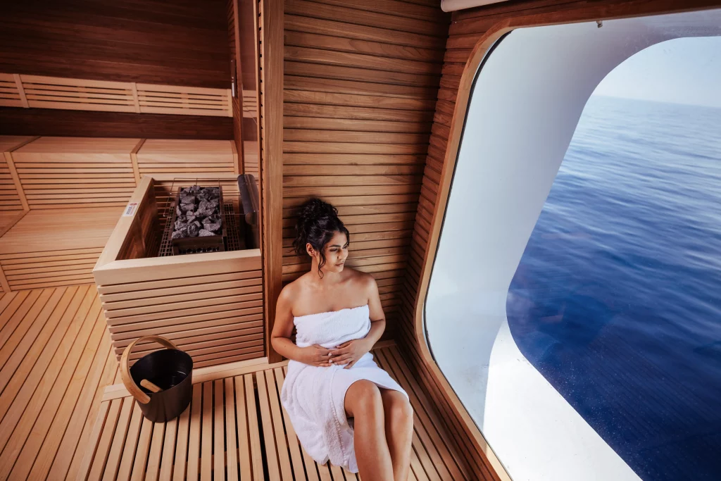 Eine Frau in einem Handtuch sitzt auf einer Holzbank in der Sauna an Bord der MSC World America und blickt während der zwei Wochen auf See durch ein großes Fenster auf das Meer.