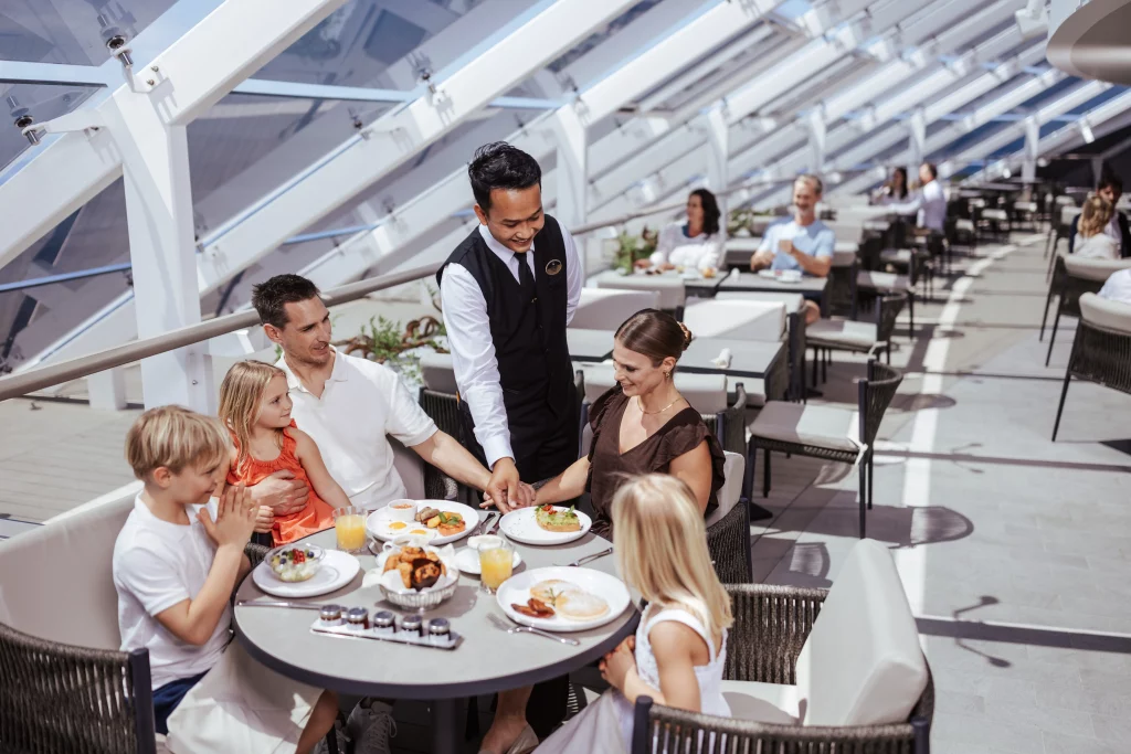 Ein Kellner serviert einer Familie mit drei Kindern das Essen an einem runden Tisch in einem hellen, modernen Restaurant mit großen Fenstern – eine Szene, die an eine Werbung für die MSC World America erinnert und sich perfekt für eine Bildproduktion eignet, die unvergessliche Momente während zweier Wochen auf See in den Mittelpunkt stellt.