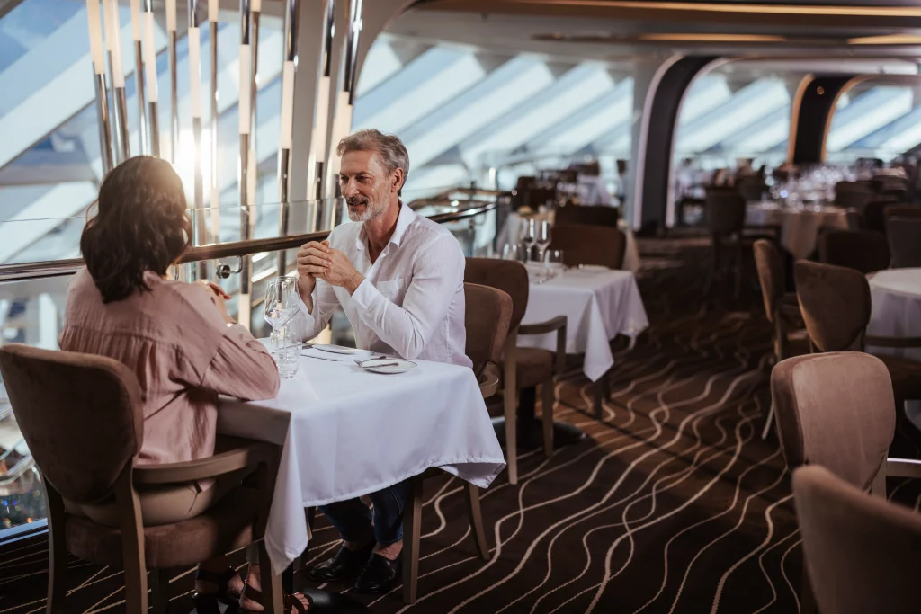 Zwei Menschen sitzen an einem Tisch in einem modernen Restaurant mit großen Fenstern an Bord der MSC World America, unterhalten sich und halten Händchen. Das Restaurant, das Teil der Imageproduktion dieses atemberaubenden Schiffes ist, ist größtenteils leer und hell beleuchtet – was vielleicht die ruhige Intimität widerspiegelt, die sich nach zwei Wochen auf See eingestellt hat.