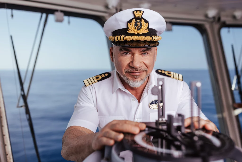 An Bord der MSC World America steht ein Schiffskapitän in Uniform am Steuer und bedient die Navigationsgeräte, während hinter ihm durch die Fenster der Ozean zu sehen ist.