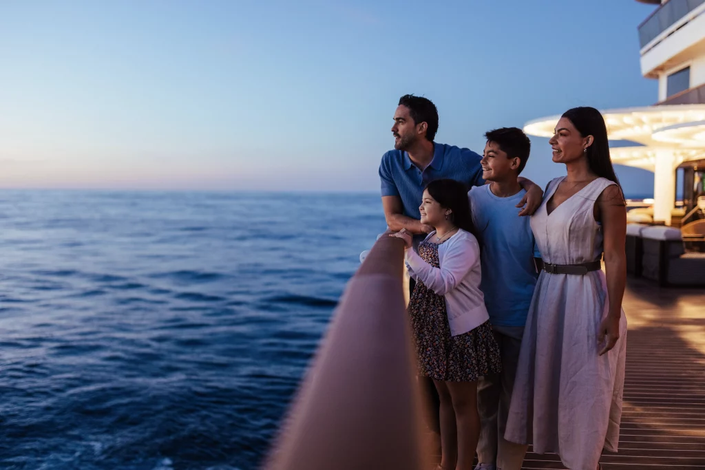 Eine vierköpfige Familie steht bei Sonnenuntergang auf dem Deck der MSC World America, blickt auf den Ozean und hält einen perfekten Moment ihrer zweiwöchigen Seereise fest.