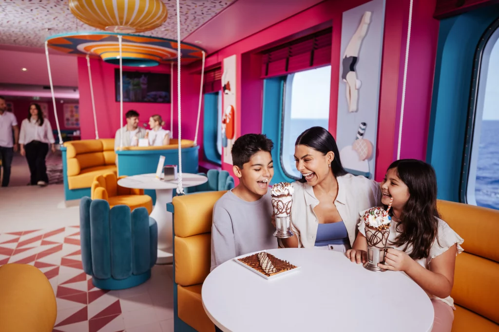 Eine Frau und zwei Kinder sitzen an einem runden Tisch in einer farbenfrohen Eisdiele an Bord der MSC World America und genießen Milchshakes und Desserts, während im Hintergrund andere Menschen zu sehen sind – der perfekte Genuss während „Zwei Wochen auf See“.