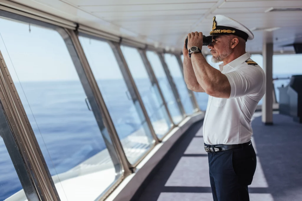 Ein Schiffskapitän in Uniform steht auf der Brücke der MSC World America und blickt durch ein Fernglas auf das Meer; im Hintergrund sind große Fenster und das blaue Meer zu sehen – eine Szene, die den Geist von „zwei Wochen auf See“ perfekt einfängt.
