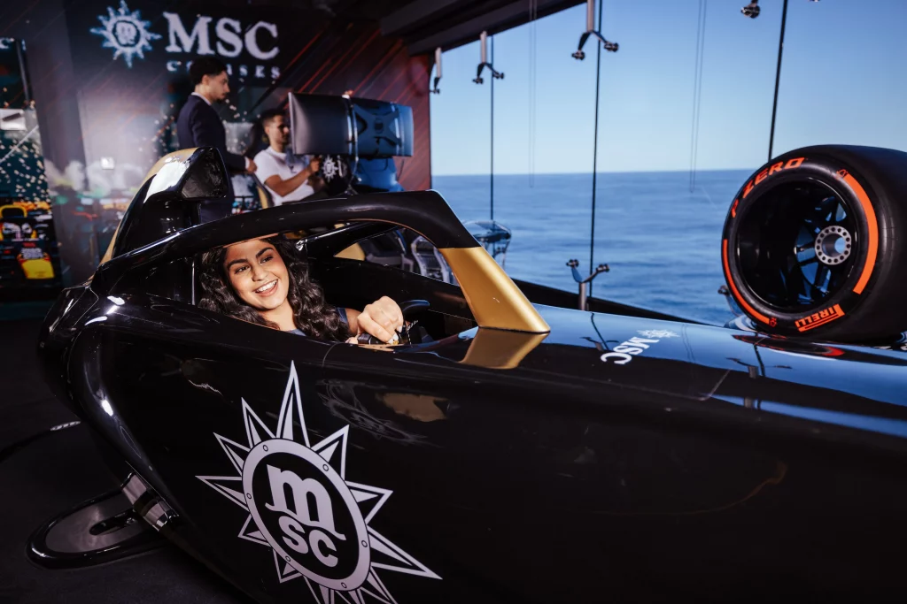 Eine Person sitzt in einem schwarzen Rennsimulator der Marke MSC, lächelt in die Kamera, im Hintergrund sind der Meerblick und technische Geräte zu sehen – ein spannender Moment der Bildproduktion während der „Zwei Wochen auf See“ an Bord der MSC World America.