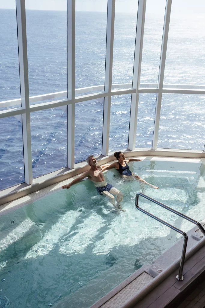 An einem sonnigen Tag an Bord der luxuriösen MSC World America entspannen sich zwei Menschen in einem Whirlpool neben großen Fenstern mit Blick auf das Meer und fangen so das perfekte „Imageproduktion“-Gefühl von zwei Wochen auf See ein.