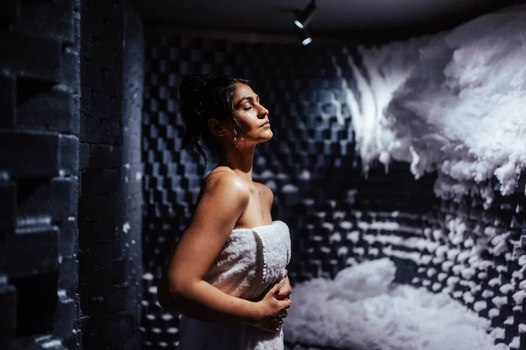 Eine in ein Handtuch gehüllte Frau steht in einem dunklen, schalldichten Raum mit Schaumstoffplatten und Kunstschnee oder Schaumstoff an den Wänden und auf dem Boden – eine Atmosphäre, die an eine immersive Bildproduktion erinnert und das surreale Gefühl von „Zwei Wochen auf See“ an Bord der MSC World America einfängt.