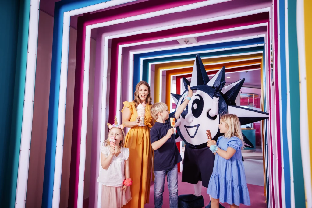 Drei Kinder und ein Erwachsener mit Eis stehen neben einer Person in einem stacheligen Sonnen-Maskottchenkostüm in einem farbenfrohen, neonbeleuchteten Tunnel – ein lebhafter Moment aus der Imageproduktion für MSC World America, der die ausgelassene Stimmung von „Zwei Wochen auf See“ einfängt.