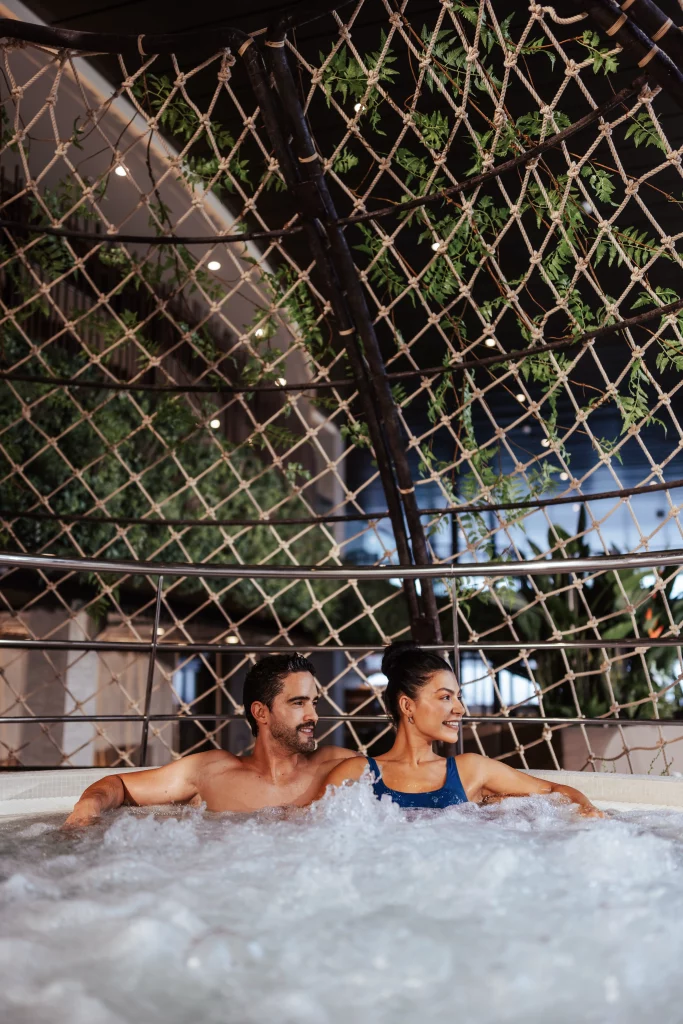 Ein Mann und eine Frau sitzen in einem sprudelnden Whirlpool, lächeln und blicken in die Ferne. Im Hintergrund sind dekorative Netzkonstruktionen und grüne Pflanzen zu sehen – ein entspannter Moment während ihrer „Zwei Wochen auf See“ an Bord der MSC World America: ein unvergessliches Erlebnis, das von Imageproduktion zum Leben erweckt wurde.