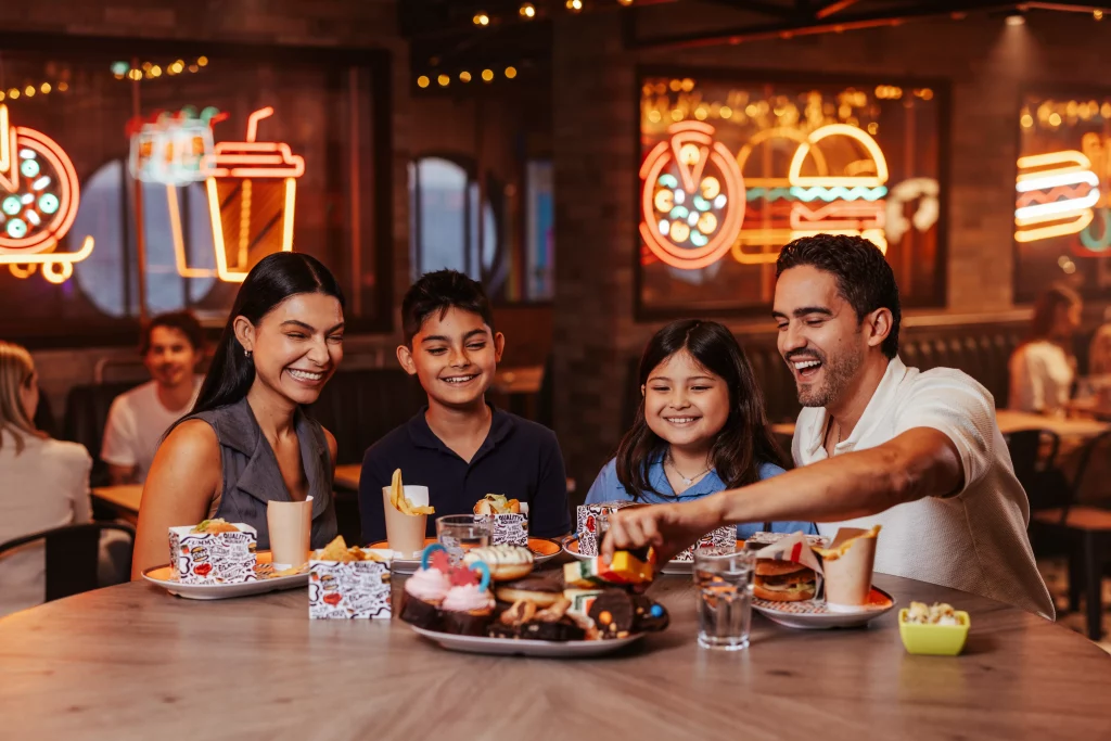 Eine vierköpfige Familie sitzt an einem runden Tisch in einem Restaurant, lächelt und isst gemeinsam, während im Hintergrund Neonreklamen für Burger und Getränke leuchten – ein Bild, das die Herzlichkeit und den Zusammenhalt einfängt, die man während der zwei Wochen auf See an Bord der MSC World America erlebt hat.