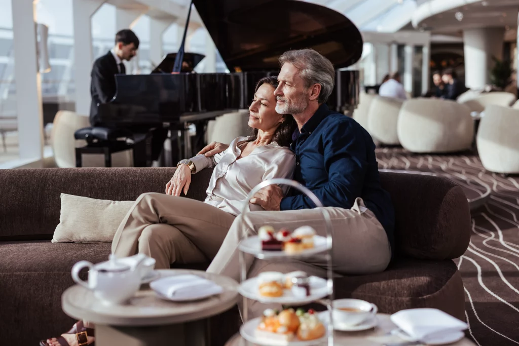 Ein Paar entspannt sich auf einem Sofa in einer modernen Lounge, auf dem Tisch stehen Tee und Gebäck, und genießt die ruhige Atmosphäre nach zwei Wochen auf See an Bord der MSC World America, während im Hintergrund ein Pianist spielt.