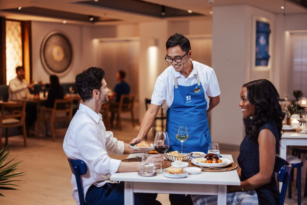 Ein Kellner in einer blauen Schürze serviert einem Mann und einer Frau, die an einem Restauranttisch an Bord der MSC World America sitzen, das Essen. Im Hintergrund sind weitere Gäste zu sehen, was eine lebhafte Szene während der „Zwei Wochen auf See“ vermittelt.