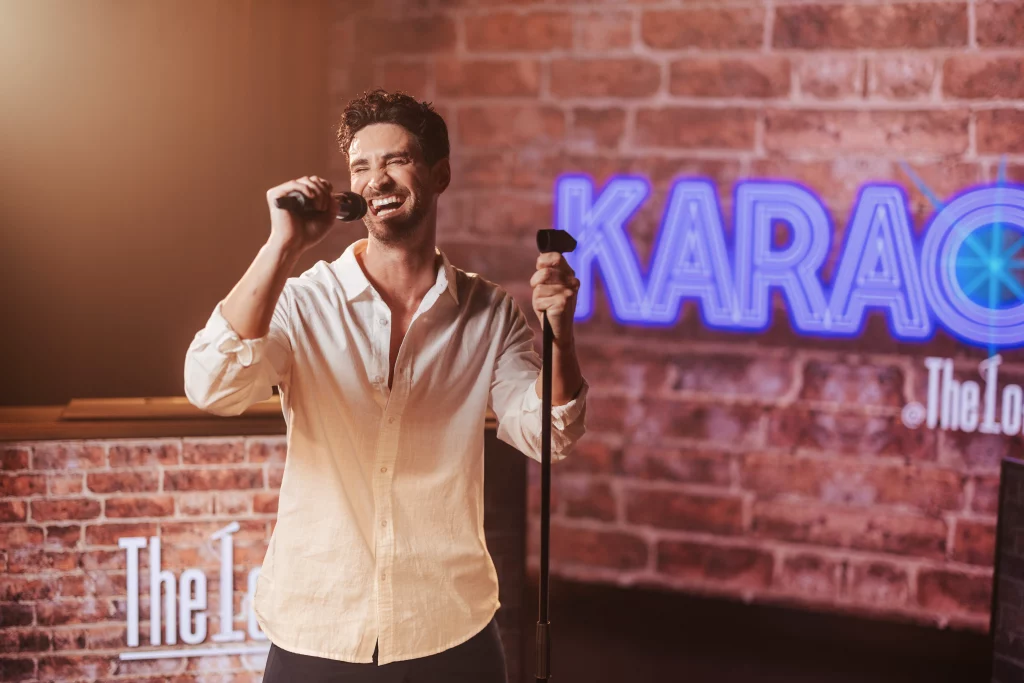 Ein Mann in einem weißen Hemd lächelt, während er auf der Bühne in ein Mikrofon singt; im Hintergrund sind ein Neon-Schild mit der Aufschrift „KARAOKE“ und eine Backsteinmauer zu sehen – ein Bild, das die lebhafte Atmosphäre einer Imageproduktion einfängt, die an Unterhaltungsabende an Bord der MSC World America erinnert.