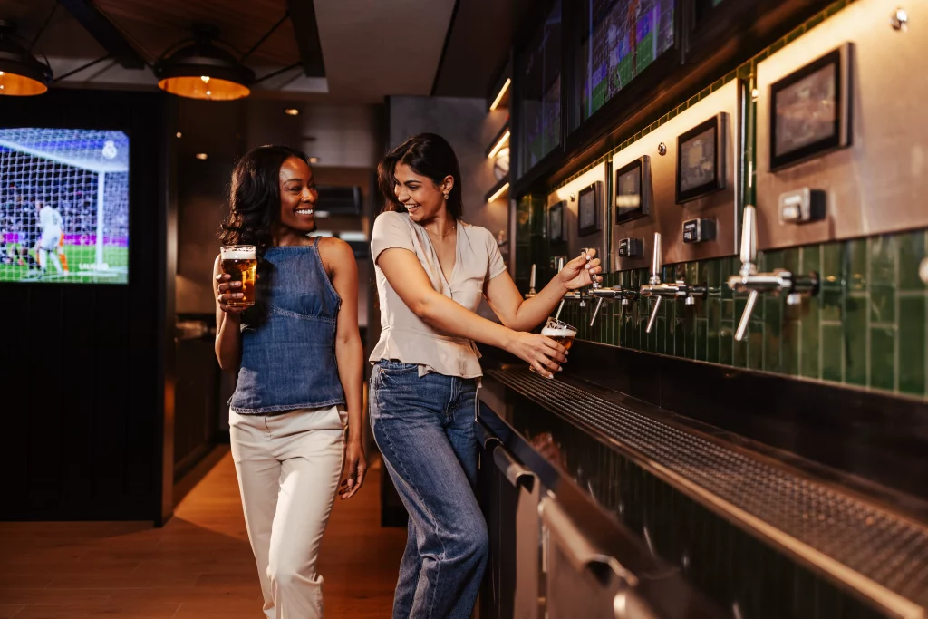 Zwei Frauen füllen an Selbstbedienungshähnen in einer Bar an Bord der MSC World America Biergläser ab, lächeln und unterhalten sich, während im Hintergrund auf einem Fernsehbildschirm ein Fußballspiel läuft – ein perfekter Moment, der im Rahmen einer Bildproduktion nach zwei Wochen auf See eingefangen wurde.