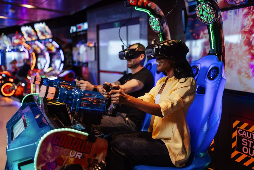 Zwei Personen mit VR-Brillen spielen ein Arcade-Shooter-Spiel, während sie in blauen Stühlen sitzen und die Spielsteuerung bedienen – in einer hell erleuchteten Spielhalle. Ein spannender Moment von Imageproduktion, vielleicht inspiriert von dem Spaß und den Abenteuern, die die Gäste nach „Zwei Wochen auf See“ an Bord der MSC World America erwarten.