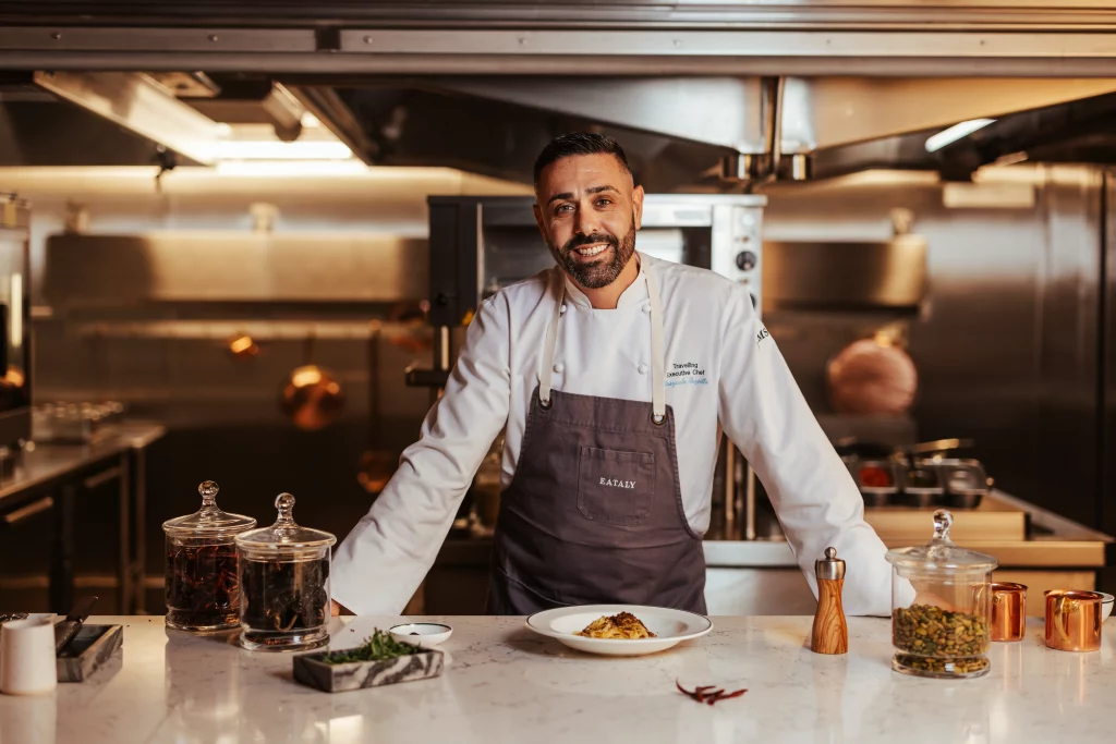 Ein Koch in weißem Kittel und Schürze steht hinter einer Theke in der Profiküche an Bord der MSC World America, vor sich ein angerichtetes Gericht, Gewürze und Kräuter, und kreiert gekonnt Aromen, die von zwei Wochen auf See inspiriert sind.