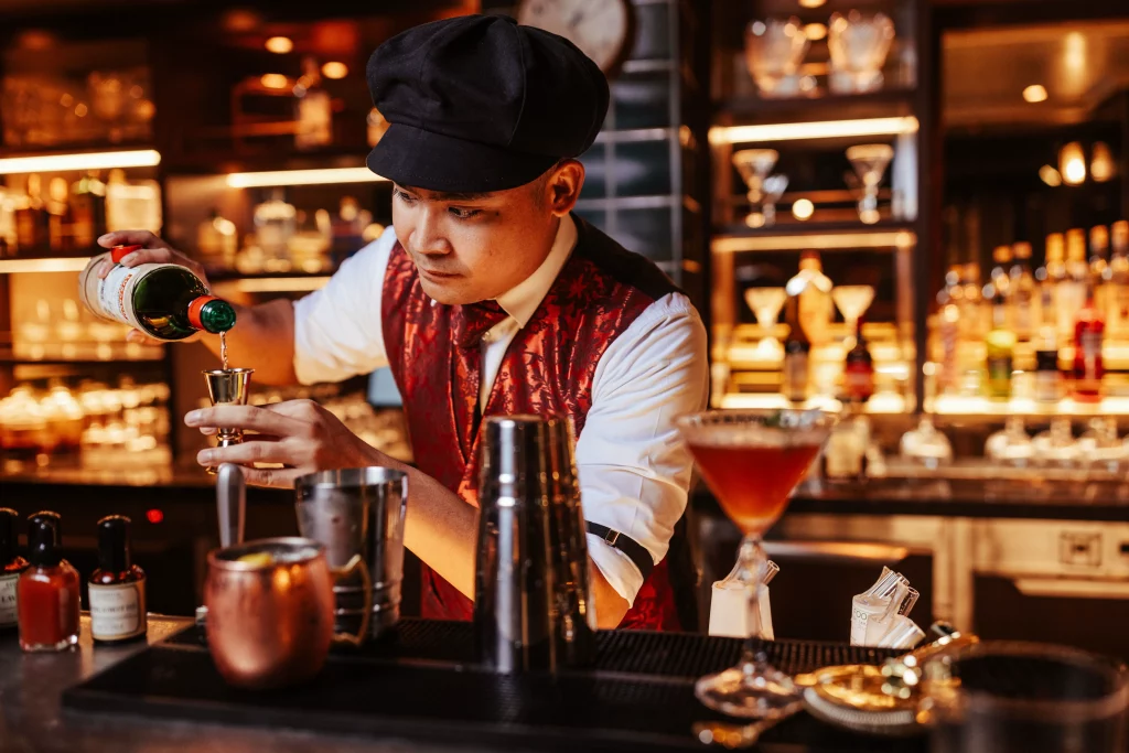 Ein Barkeeper in einer roten Weste schenkt an einer Bar, umgeben von Cocktailgläsern, Shakers und Flaschen, Spirituosen in einen Jigger ein – und fängt damit die lebhafte Atmosphäre der MSC World America während einer Imageproduktion nach zwei Wochen auf See ein.