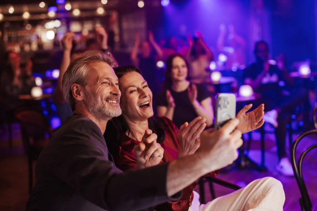 Ein Mann und eine Frau lächeln und machen ein Selfie, während sie im Publikum eines lebhaften, schwach beleuchteten Veranstaltungsortes sitzen, und halten so die Aufregung ihres MSC World America-Abenteuers während zwei Wochen auf See fest.
