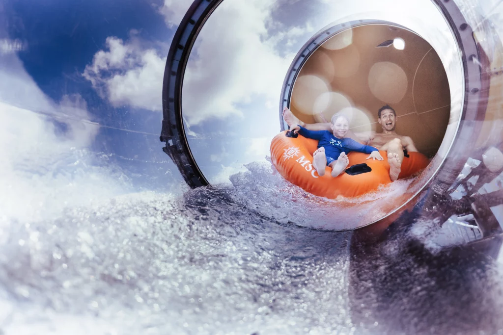 Zwei Personen rutschen an Bord der MSC World America auf einem großen orangefarbenen Schlauchboot eine kurvenreiche Wasserrutsche hinunter und landen mit einem Spritzer im Wasser, wobei durch die transparenten Abschnitte der Rutsche Wolken und blauer Himmel zu sehen sind – perfekt eingefangen während einer Bildproduktion für „Zwei Wochen auf See“.